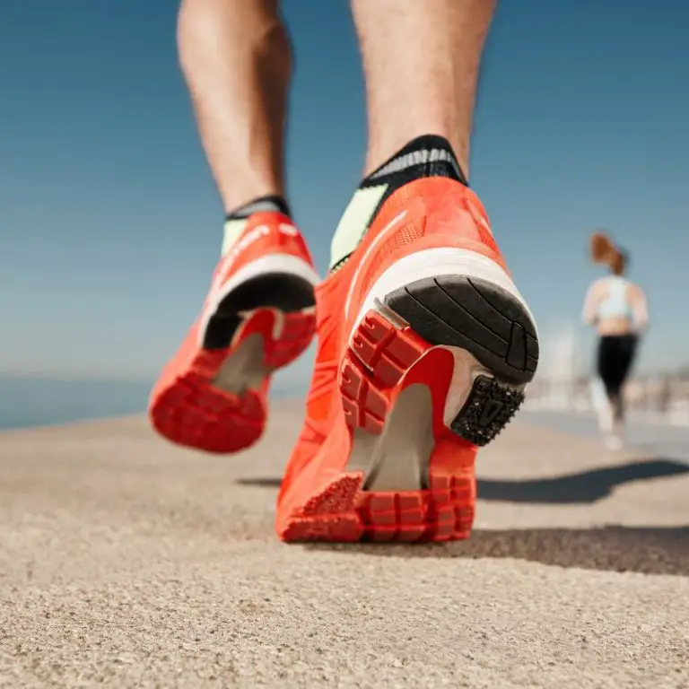 Close-up of a person running in bright orange running shoes on a sunny day, with a blurred runner in the background